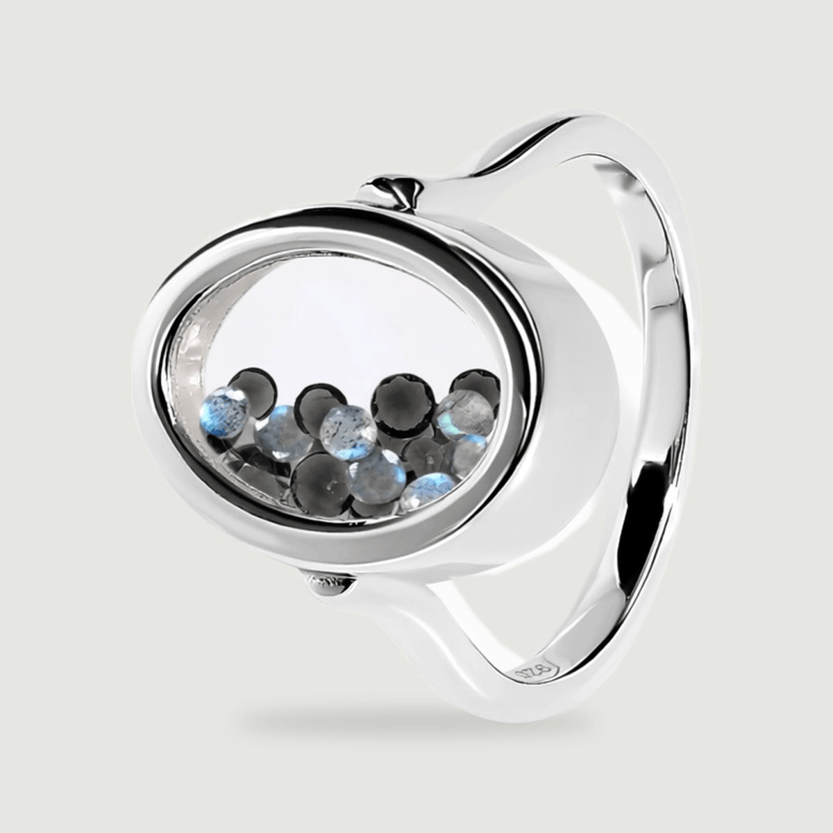 An angled sterling silver ring reveals its oval chamber, reflective band, and small round black and blue gray gems.