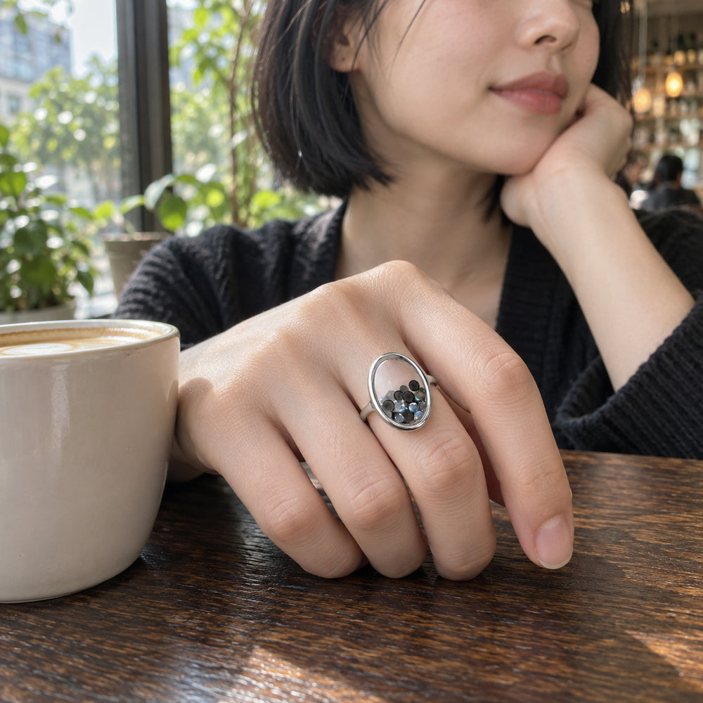 A sterling silver ring with black and blue gray gems is worn on a hand resting beside a coffee cup in a cafe.