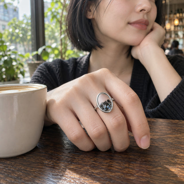 A sterling silver ring with black and blue gray gems is worn on a hand resting beside a coffee cup in a cafe.
