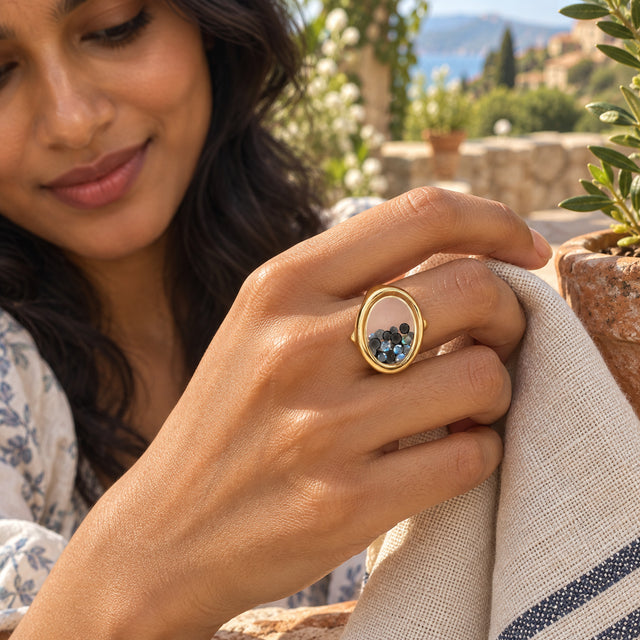 A yellow gold vermeil ring with a clear oval chamber is worn on a hand holding a linen napkin on a garden terrace.