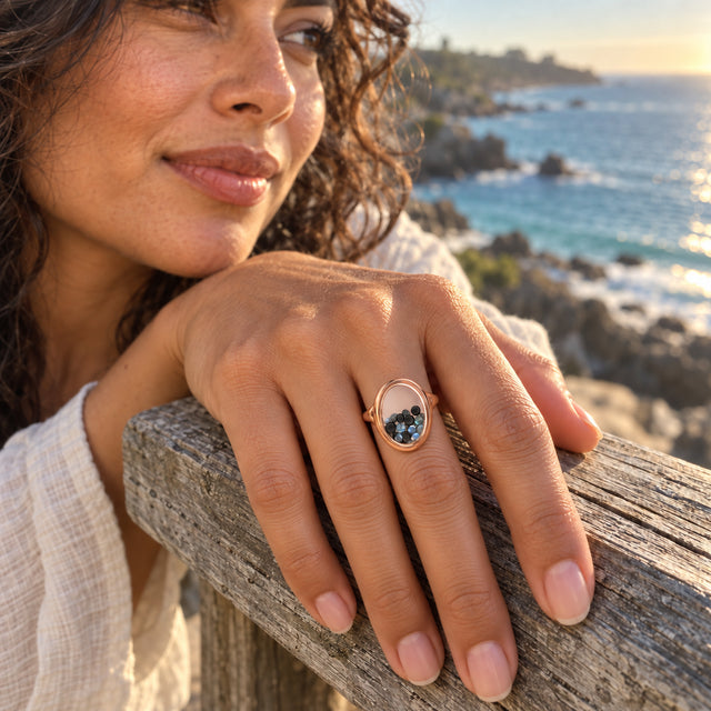A rose gold vermeil ring containing small dark and iridescent gems is worn on a hand resting on a wooden seaside railing.