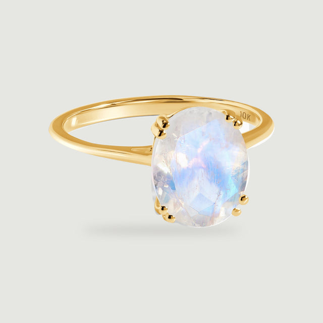 A faceted oval moonstone rises above a slim yellow gold band on a light gray surface.