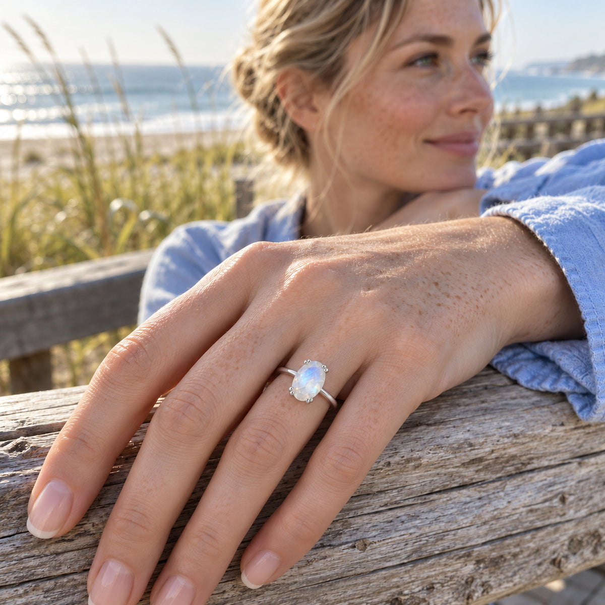 A white gold moonstone ring is worn on a freckled hand resting on weathered wood beside the sea.