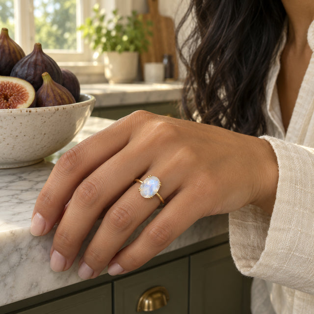 A yellow gold moonstone ring is shown on a hand resting on a marble kitchen counter beside a bowl of figs.