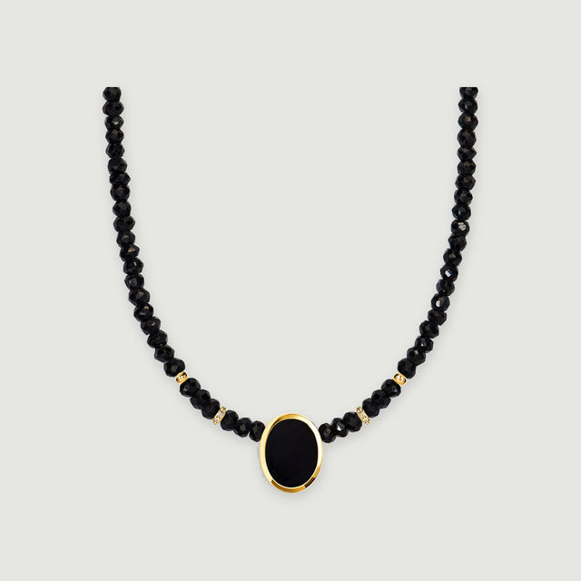 A black faceted bead necklace with an oval black pendant and yellow gold bezel is centered on a light gray background.