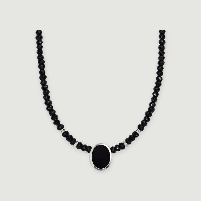 A strand of faceted black beads curves around an oval black pendant in a polished silver setting on light gray.
