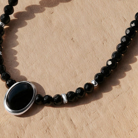 A silver bezel frames the oval black stone beside black beads and small silver spacers on light wood.