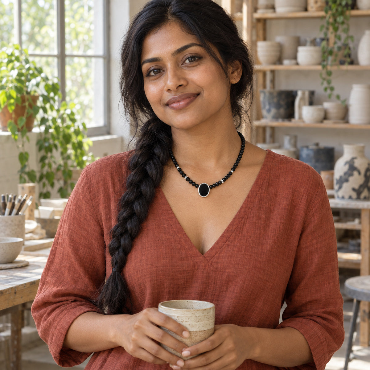 A woman with a long dark braid wears the black bead necklace with a silver oval bezel in a pottery workshop.