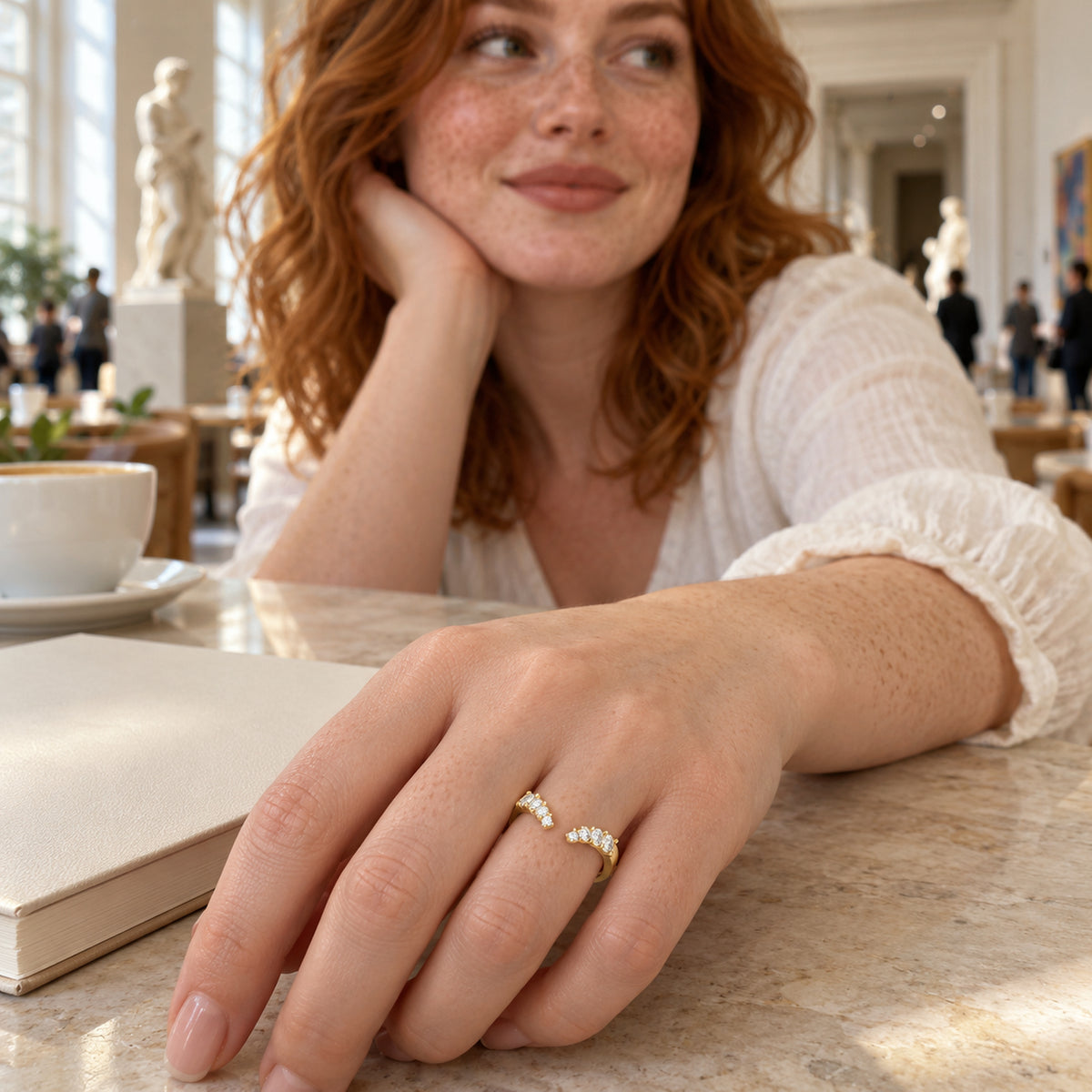 A yellow gold vermeil ring with an open front is shown on a freckled hand beside a notebook in a museum cafe.