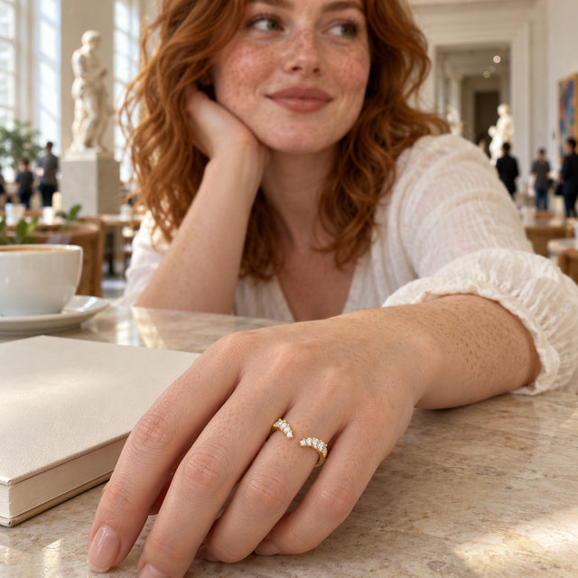A yellow gold vermeil ring with an open front is shown on a freckled hand beside a notebook in a museum cafe.