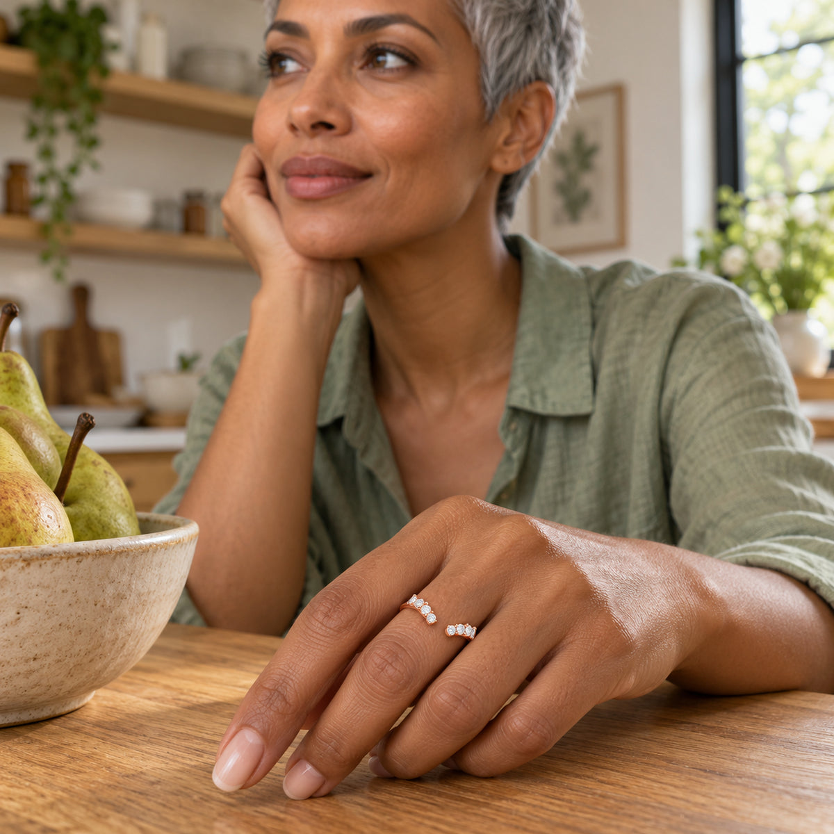 A rose gold vermeil band with clear round stones is worn on a hand resting on a wooden kitchen counter.