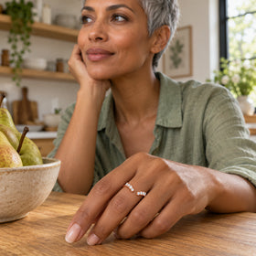 A rose gold vermeil band with clear round stones is worn on a hand resting on a wooden kitchen counter.