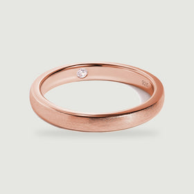 A rose gold ring rests at an angle with a brushed band and a clear stone set into its inner surface.