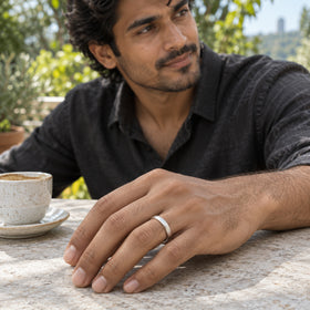 A silver band is worn on a man's hand resting beside a ceramic coffee cup on an outdoor stone table.