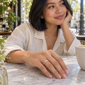 A woman wears a rose gold band with her hand resting on a marble cafe table beside a white cup.