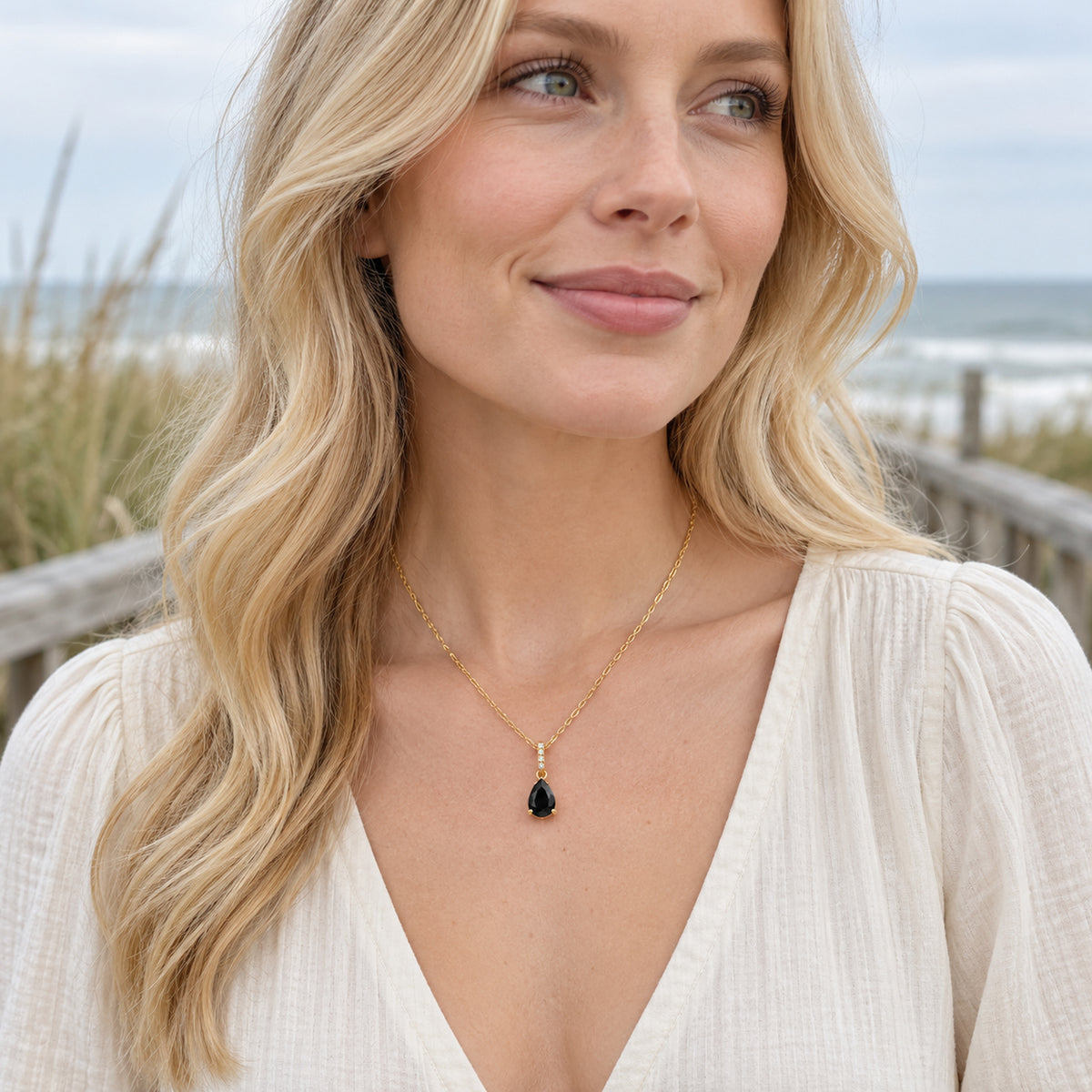 A blonde woman wears a yellow gold vermeil black onyx necklace above a cream blouse on a coastal boardwalk.
