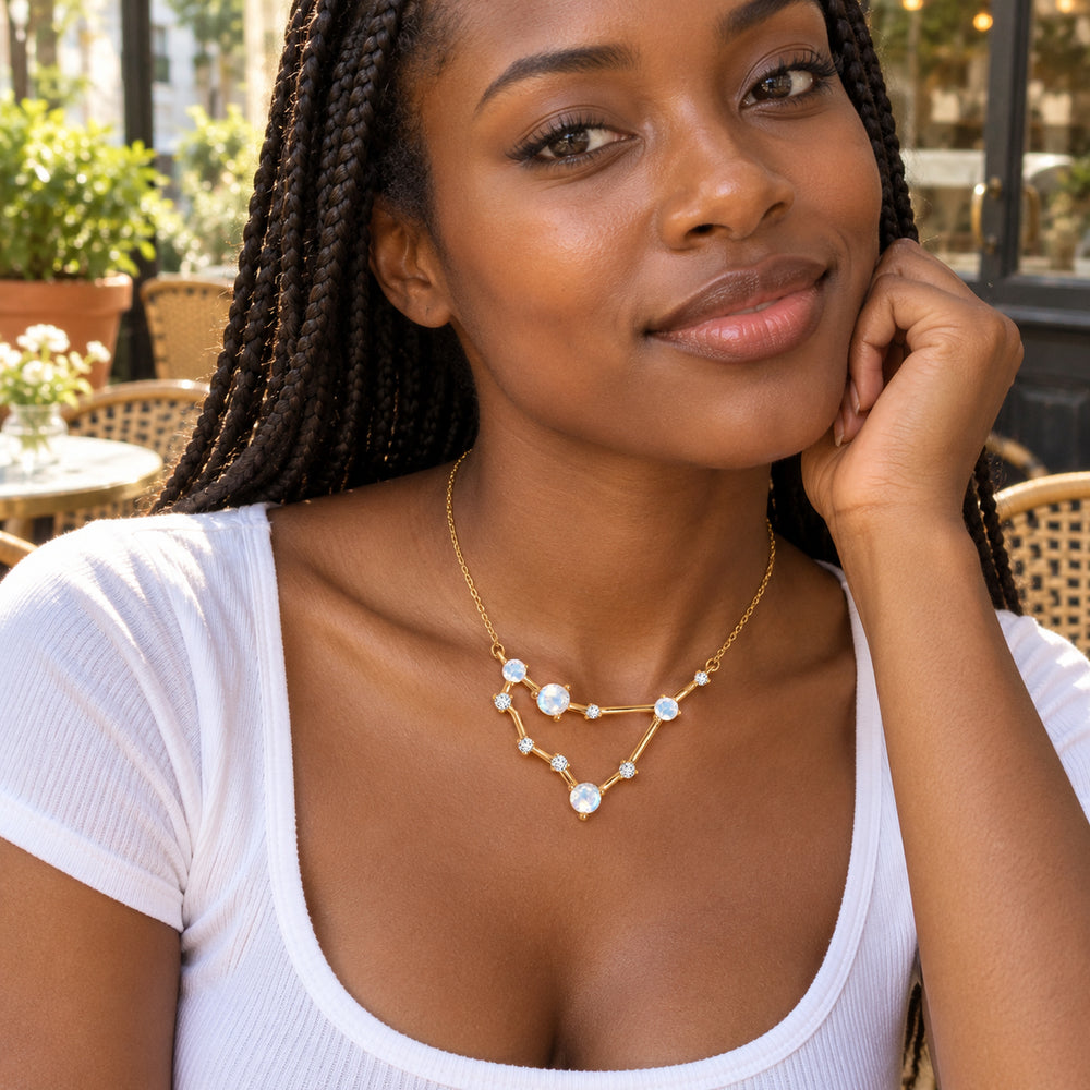 A woman with braided hair wears the yellow gold necklace with a white scoop-neck top at an outdoor cafe.