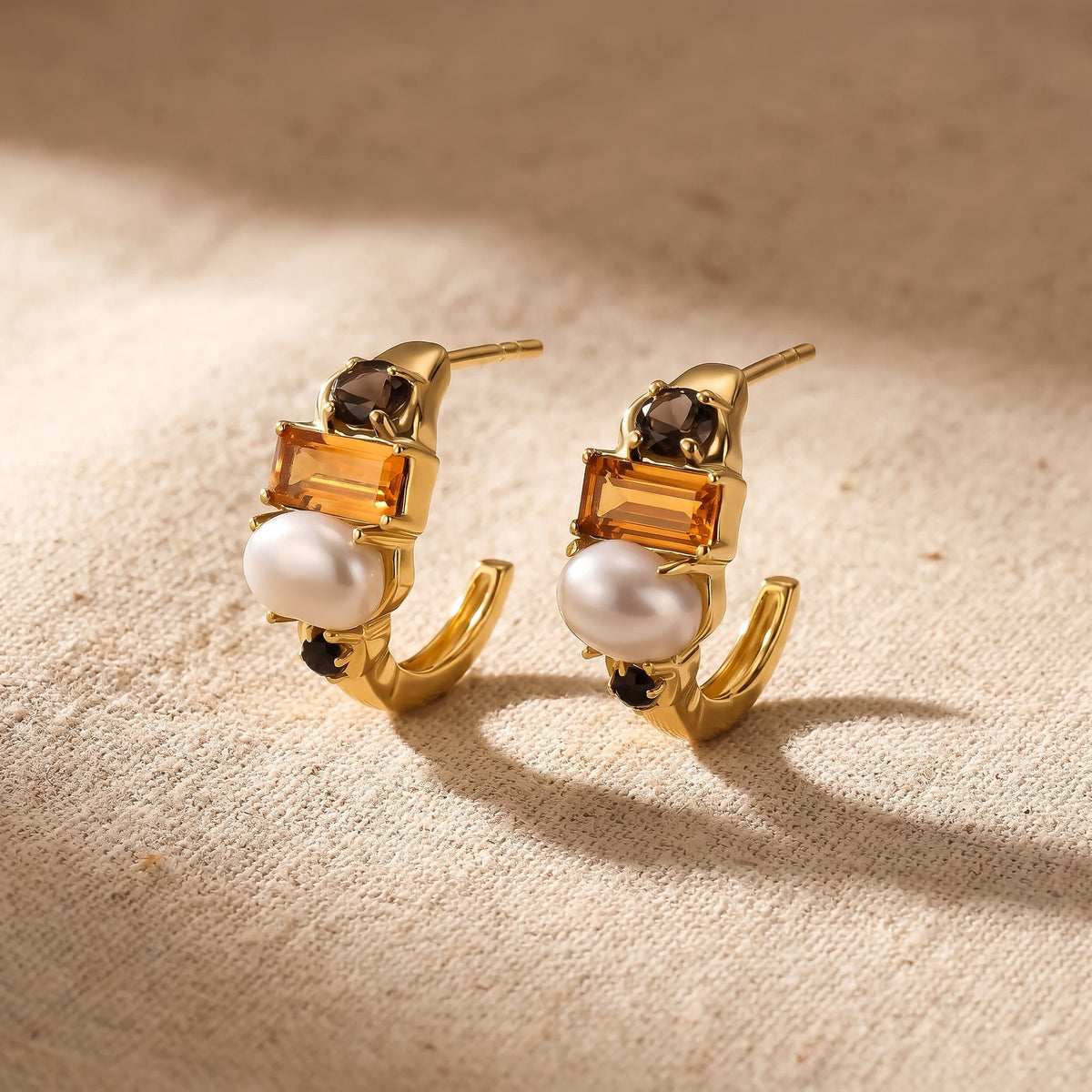 Two gold-tone pearl and citrine hoops rest at an angle on textured beige fabric.