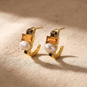 Two gold-tone pearl and citrine hoops rest at an angle on textured beige fabric.