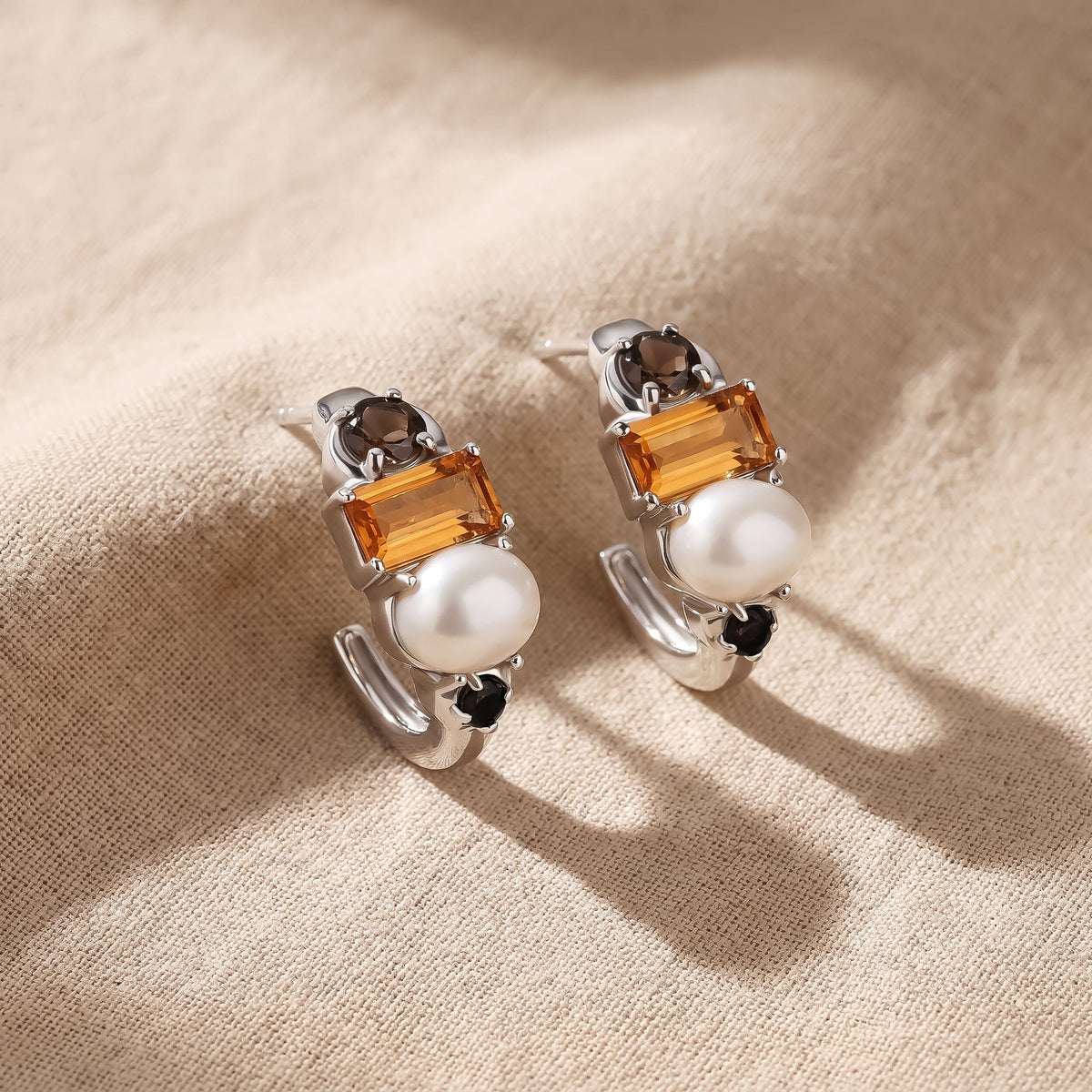 Silver-tone earrings lie side by side on beige cloth with their orange stones and white pearls facing upward.