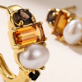 A close view reveals the gold-tone prongs around an orange rectangular stone, a white pearl, and dark round accents.