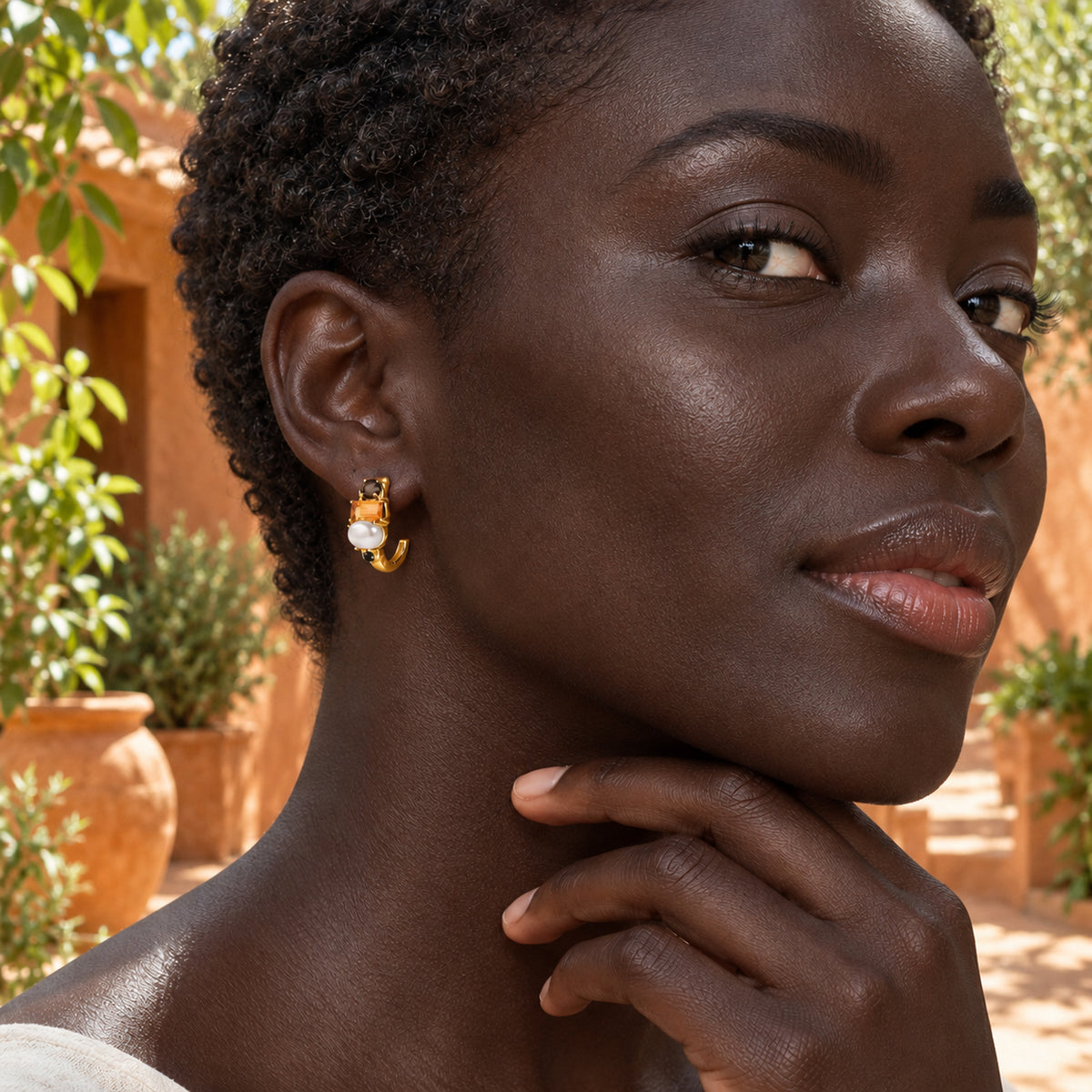 A woman with short curls wears a gold-tone pearl and orange-stone hoop beside terracotta walls and green plants.