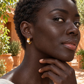 A woman with short curls wears a gold-tone pearl and orange-stone hoop beside terracotta walls and green plants.