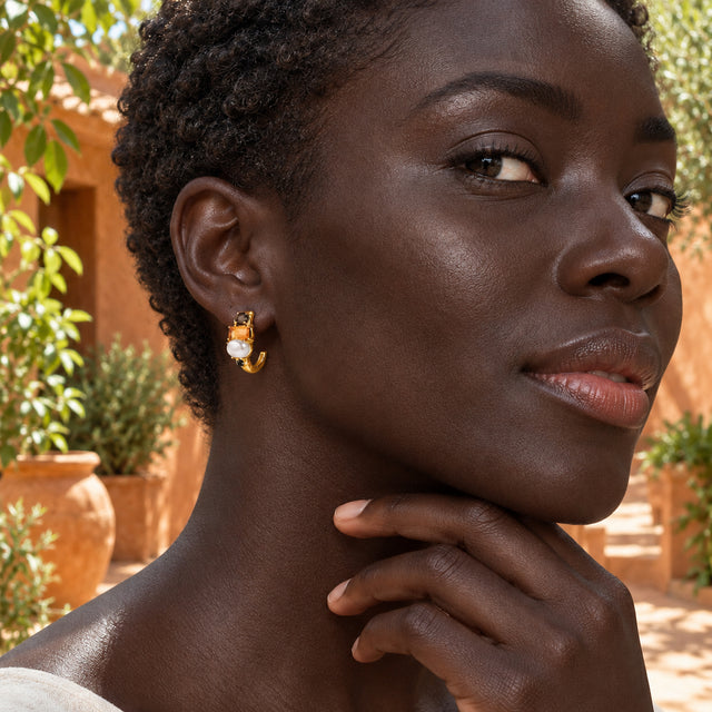 A woman with short curls wears a gold-tone pearl and orange-stone hoop beside terracotta walls and green plants.