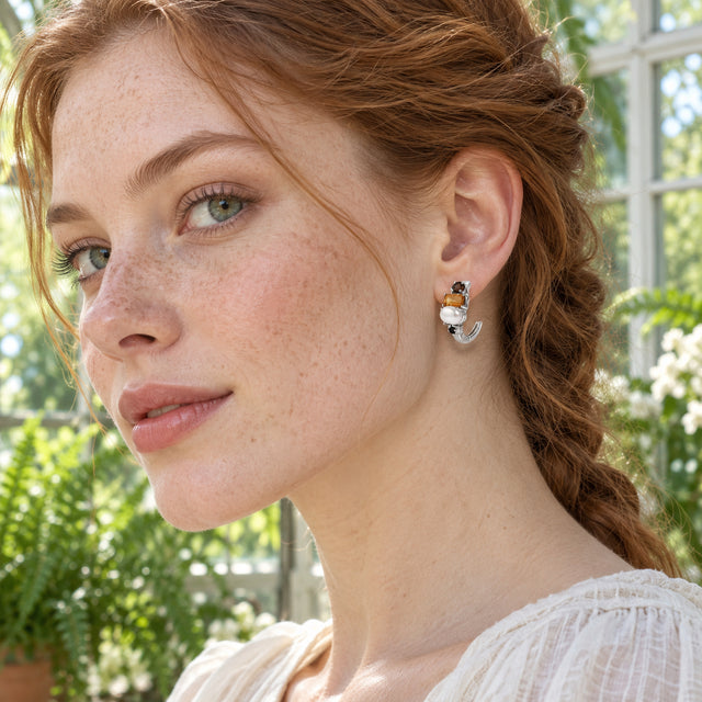 A freckled woman with auburn hair wears a silver-tone hoop with a white pearl and an orange rectangular stone in a sunlit conservatory.