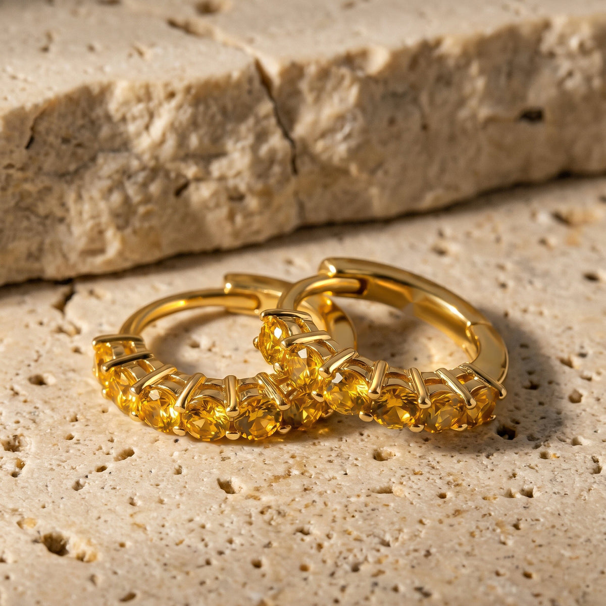 Two gold-tone citrine hoops lie together on a textured beige stone tabletop.