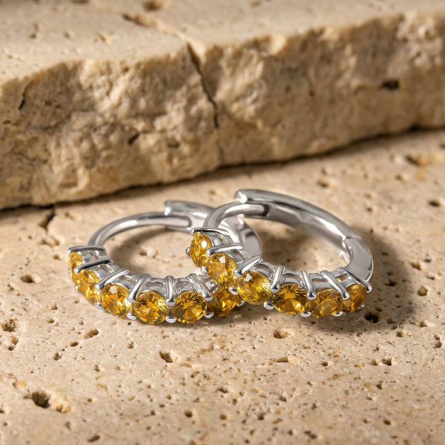 A pair of silver-tone citrine hoops displays its amber stones on a pale stone surface.