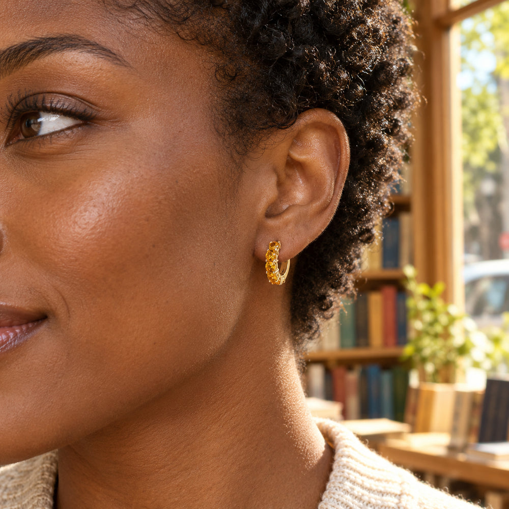 A woman with short natural curls wears a small gold-tone citrine hoop beside a sunlit bookshop window.
