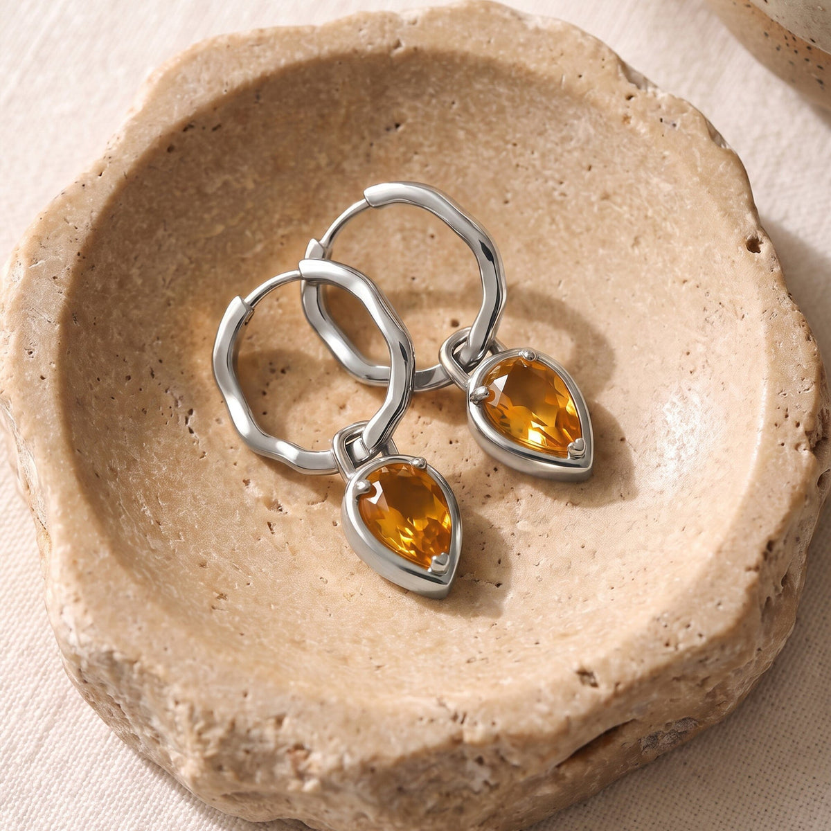 White rhodium plated hoops and amber citrine drops are arranged on a warm stone dish.