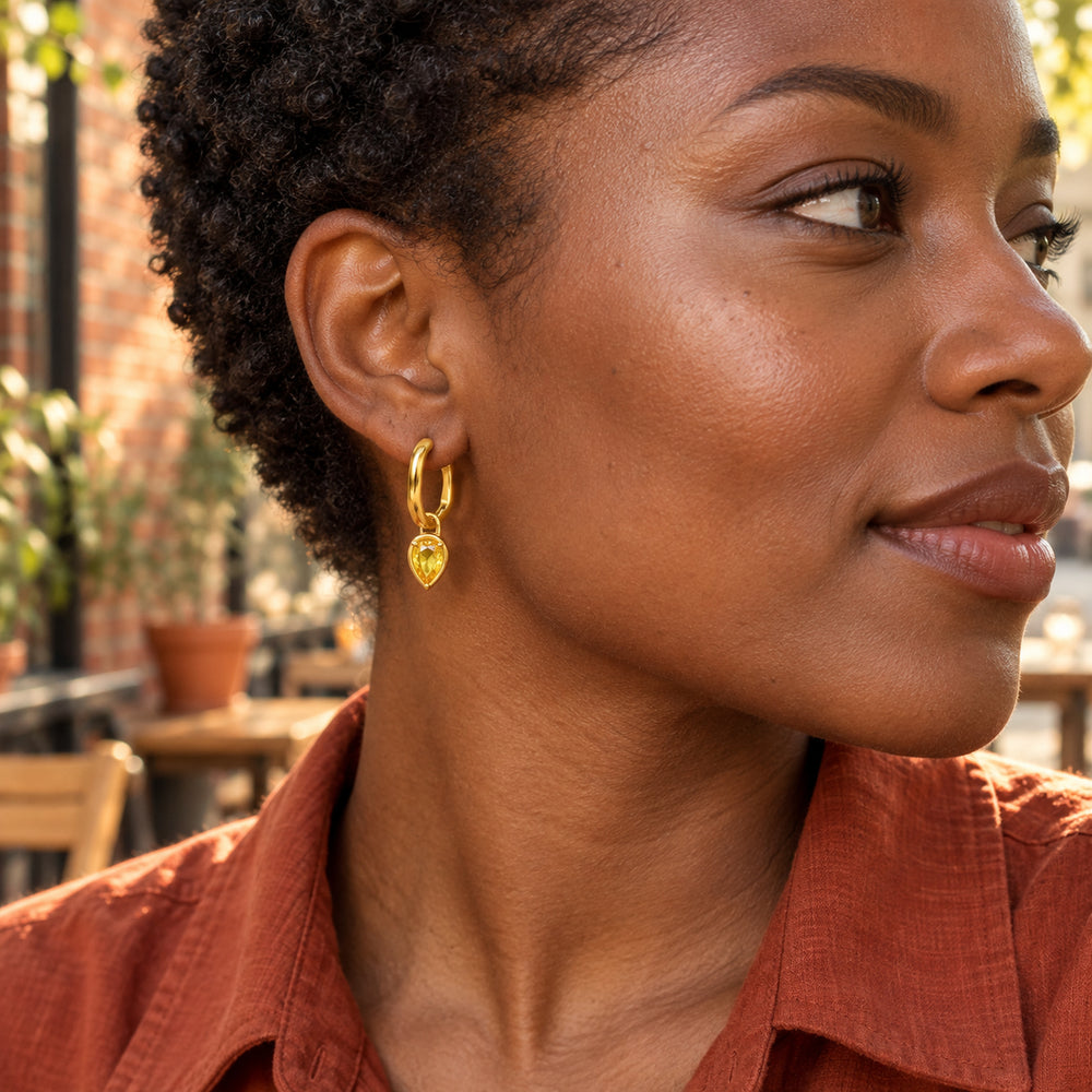 A woman with short coiled hair wears a yellow gold plated citrine hoop beside a sunlit brick cafe.