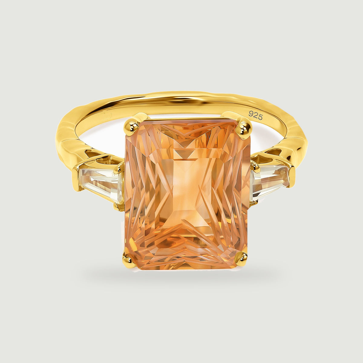 A rectangular amber citrine and two clear side stones sit in a yellow gold ring on a light gray surface.