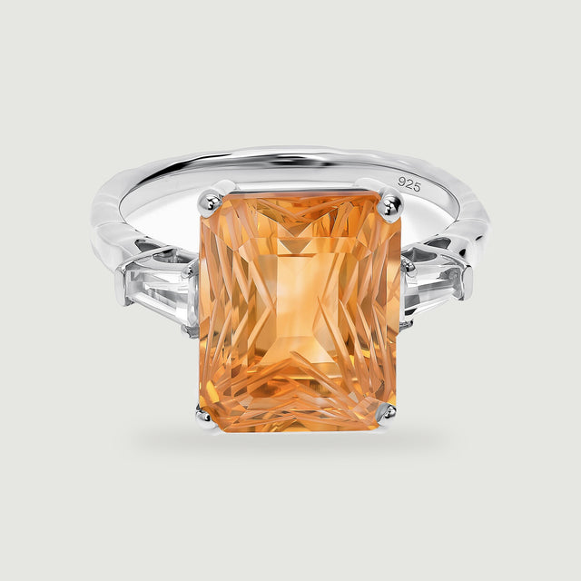 A silver ring with a carved rectangular citrine is centered above a soft shadow on a light gray background.