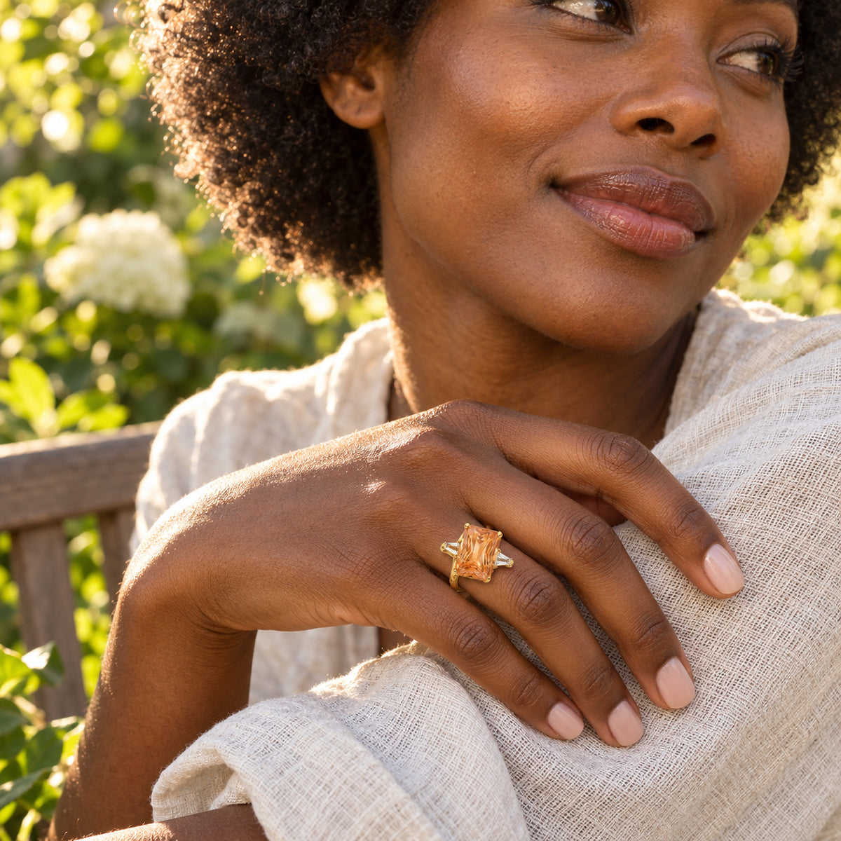 A woman with curly dark hair wears a gold citrine ring while resting her hand on cream linen in a leafy garden.