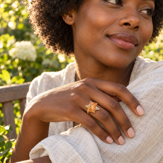 A woman with curly dark hair wears a gold citrine ring while resting her hand on cream linen in a leafy garden.