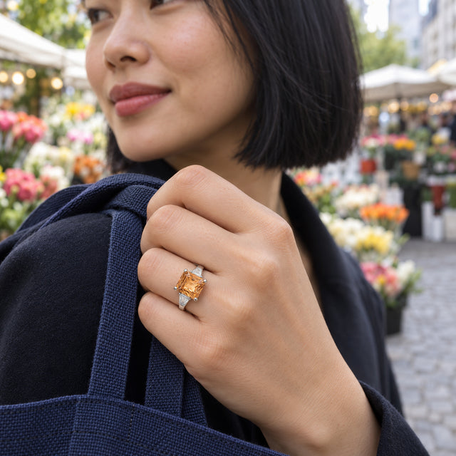 A silver citrine ring is shown on a woman holding a navy bag strap beside outdoor flower stalls.