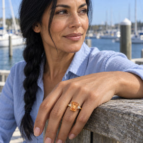 A woman with a long dark braid wears a gold citrine ring while resting her hand on a wooden railing beside sailboats.