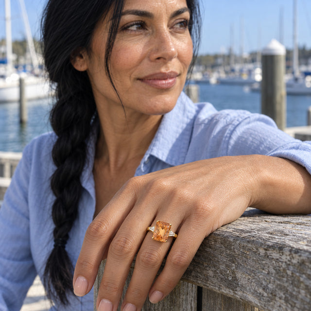 A woman with a long dark braid wears a gold citrine ring while resting her hand on a wooden railing beside sailboats.