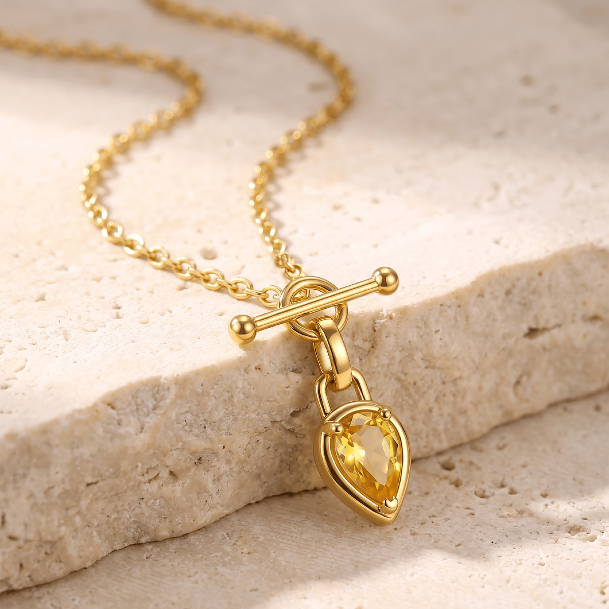 The gold toned necklace drapes across pale textured stone with the yellow pendant resting at the edge.