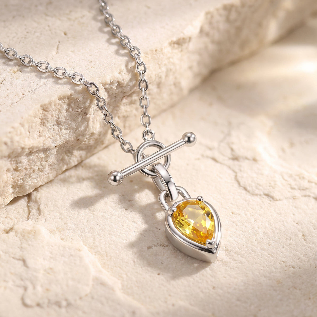 A white rhodium plated chain lies on a pale stone surface beside its pear shaped yellow pendant.