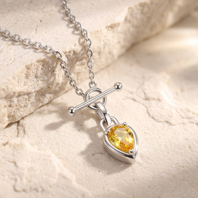 A white rhodium plated chain lies on a pale stone surface beside its pear shaped yellow pendant.