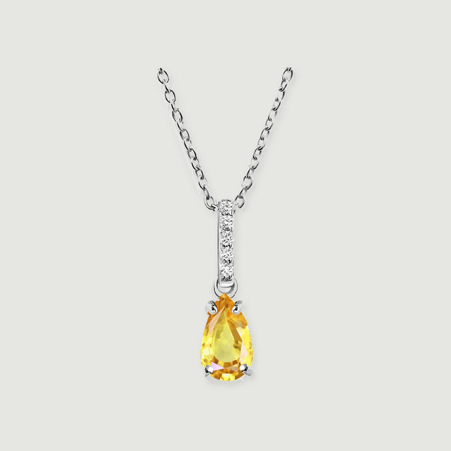 A silver necklace displays a yellow pear-shaped pendant beneath five round clear stones on a pale gray background.