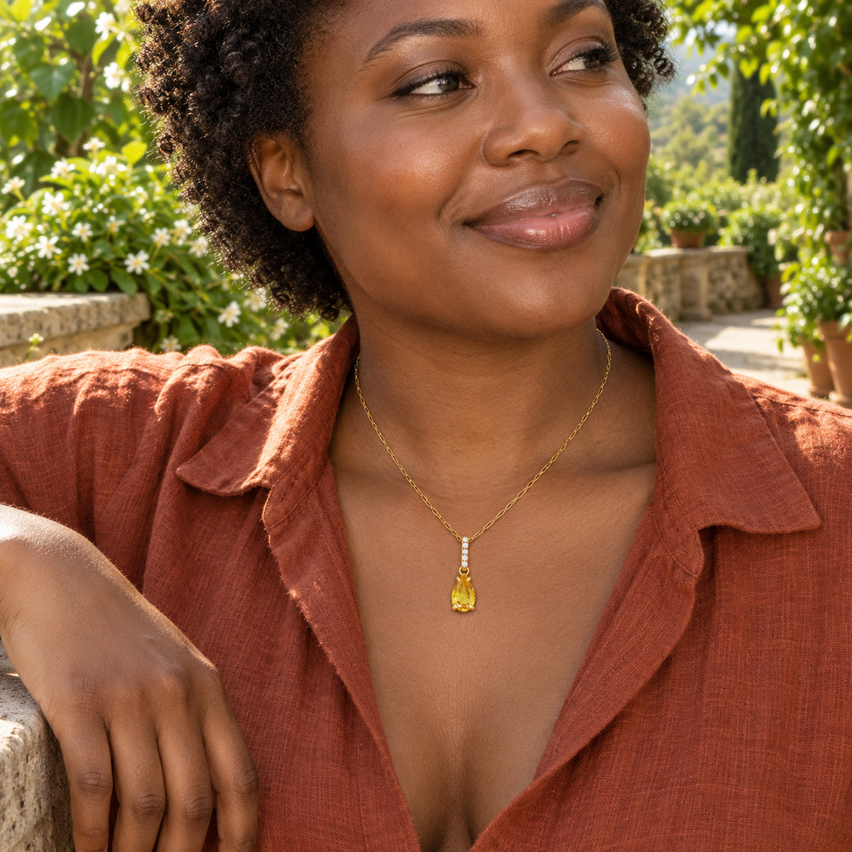A woman in a rust linen blouse wears a gold necklace with a yellow pear-shaped pendant on a leafy terrace.