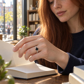 A woman with auburn hair wears a silver ring with a black pear-shaped stone while turning a book page beside a sunny window.