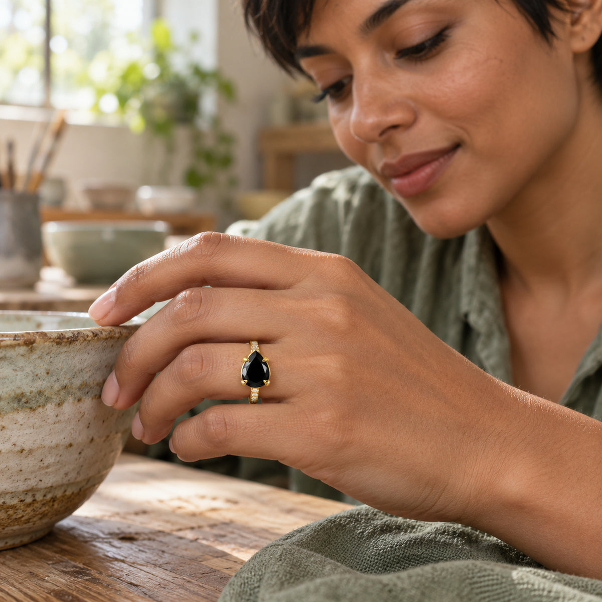 A woman with short dark hair wears a yellow gold ring with a black pear-shaped stone beside a speckled ceramic bowl in a pottery studio.