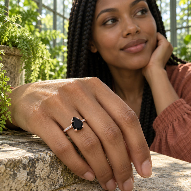 A woman with braided hair wears a rose gold ring with a black faceted stone as her hand rests on a stone ledge among green plants.