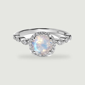 A white gold ring with a round faceted moonstone, a diamond halo and scalloped diamond shoulders rests on light gray.